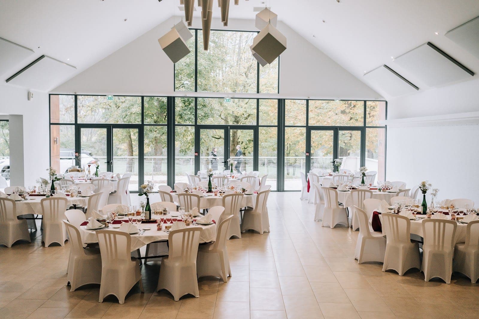 Reception room with round tables set for a wedding, overlooking trees through large windows.