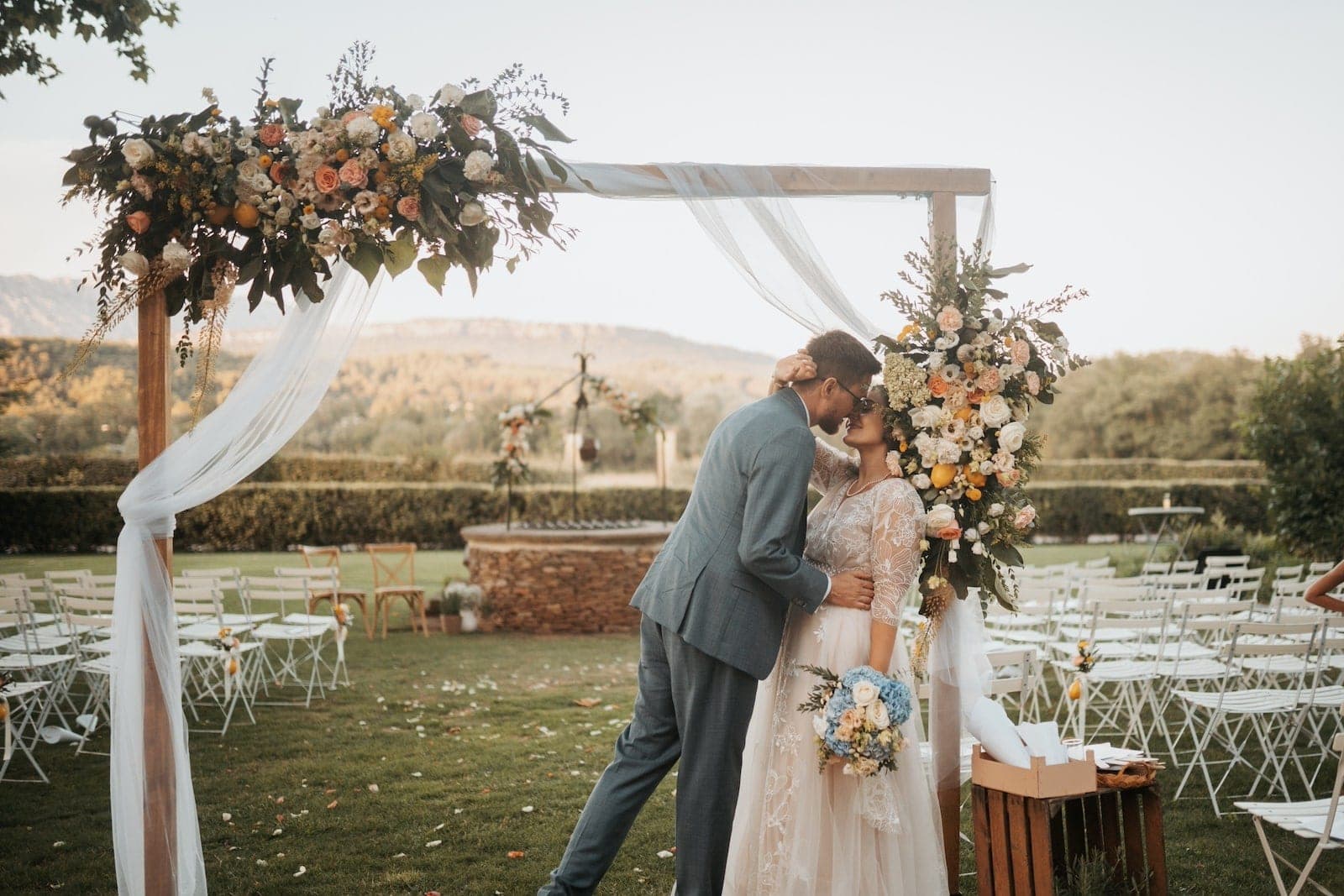 A couple kisses under a floral archway set up for a wedding ceremony in a garden.