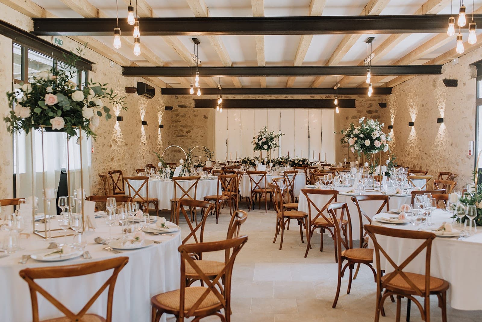 A wedding reception room with round tables set for dinner, featuring floral centerpieces and wooden chairs.