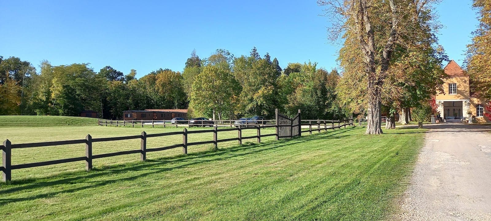 A sunny day shows a large green lawn with a wooden fence and trees, leading to a building.