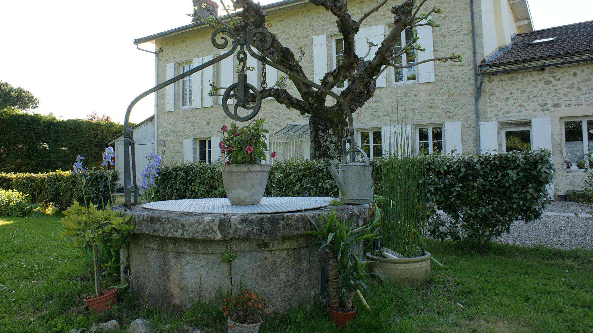 A stone well with a metal pulley system in a garden with a stone house behind.