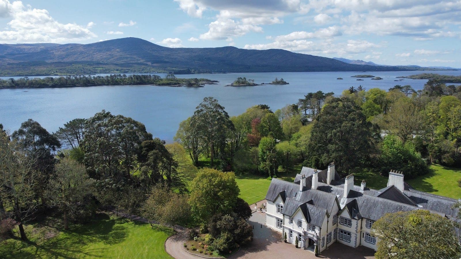 A large white manor house with grey roofs sits on green lawns beside a lake with islands and mountains beyond.