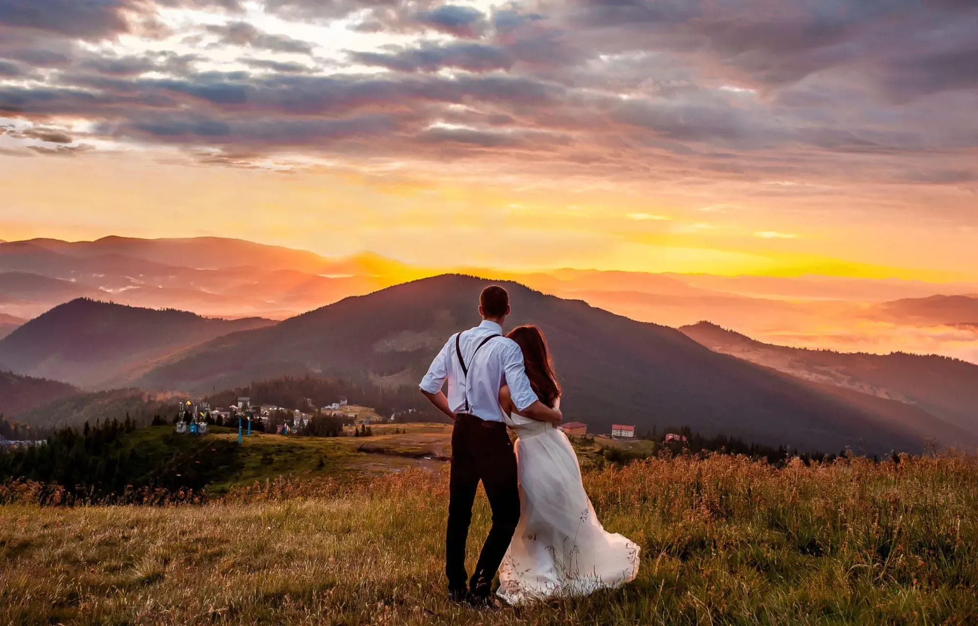 Couple embracing on a grassy hill overlooking mountains at sunset.