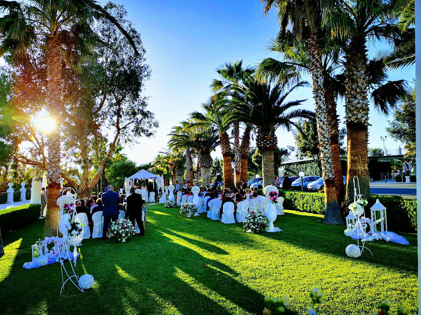 Wedding ceremony setup on a green lawn with palm trees and white chairs.