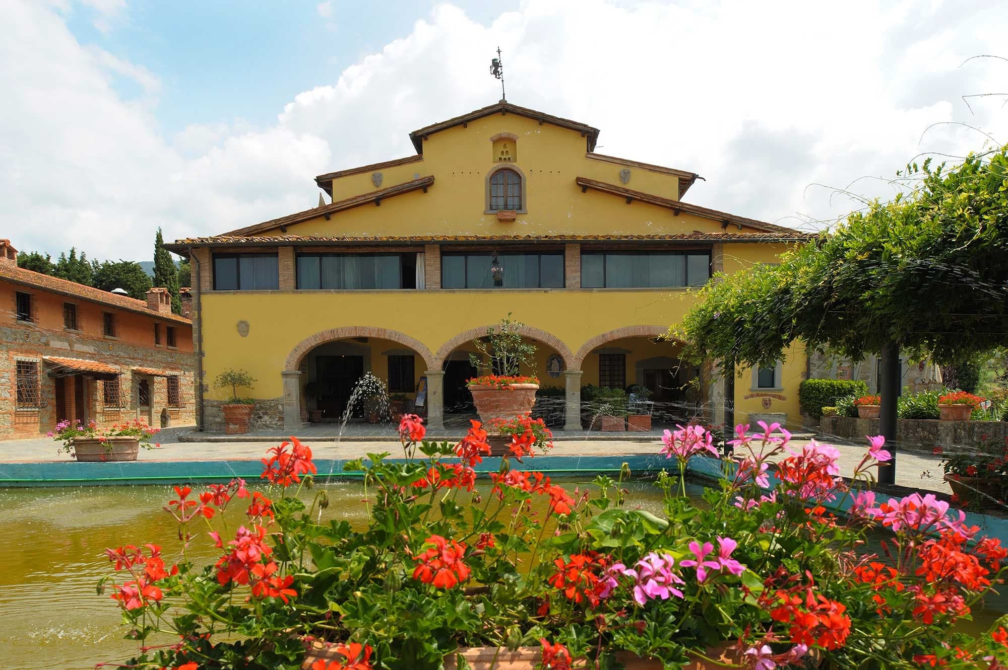 Yellow building with arched walkways and a fountain in front, surrounded by flowers.