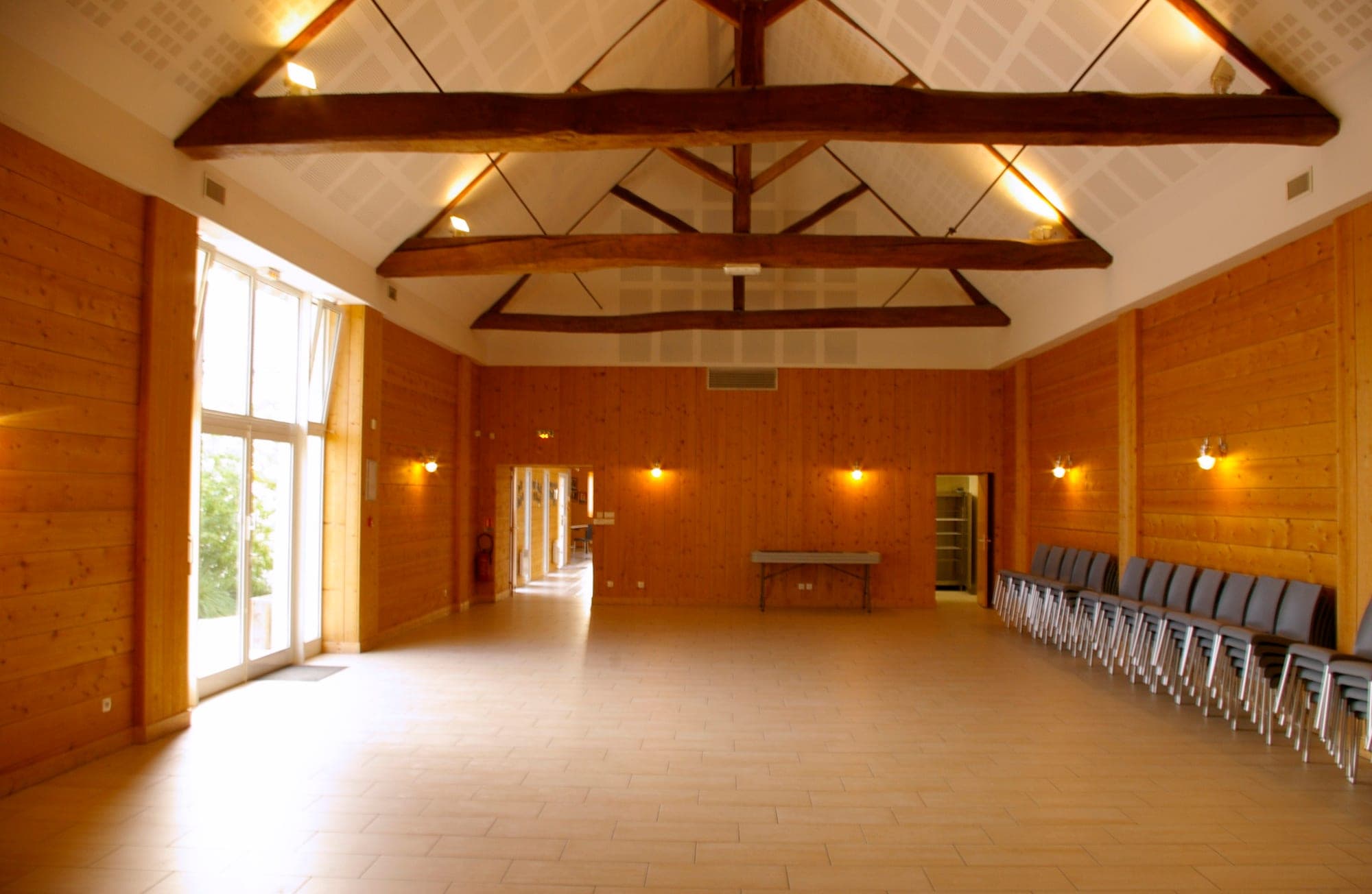 Empty reception hall with wooden walls, chairs stacked on the side, and large windows.