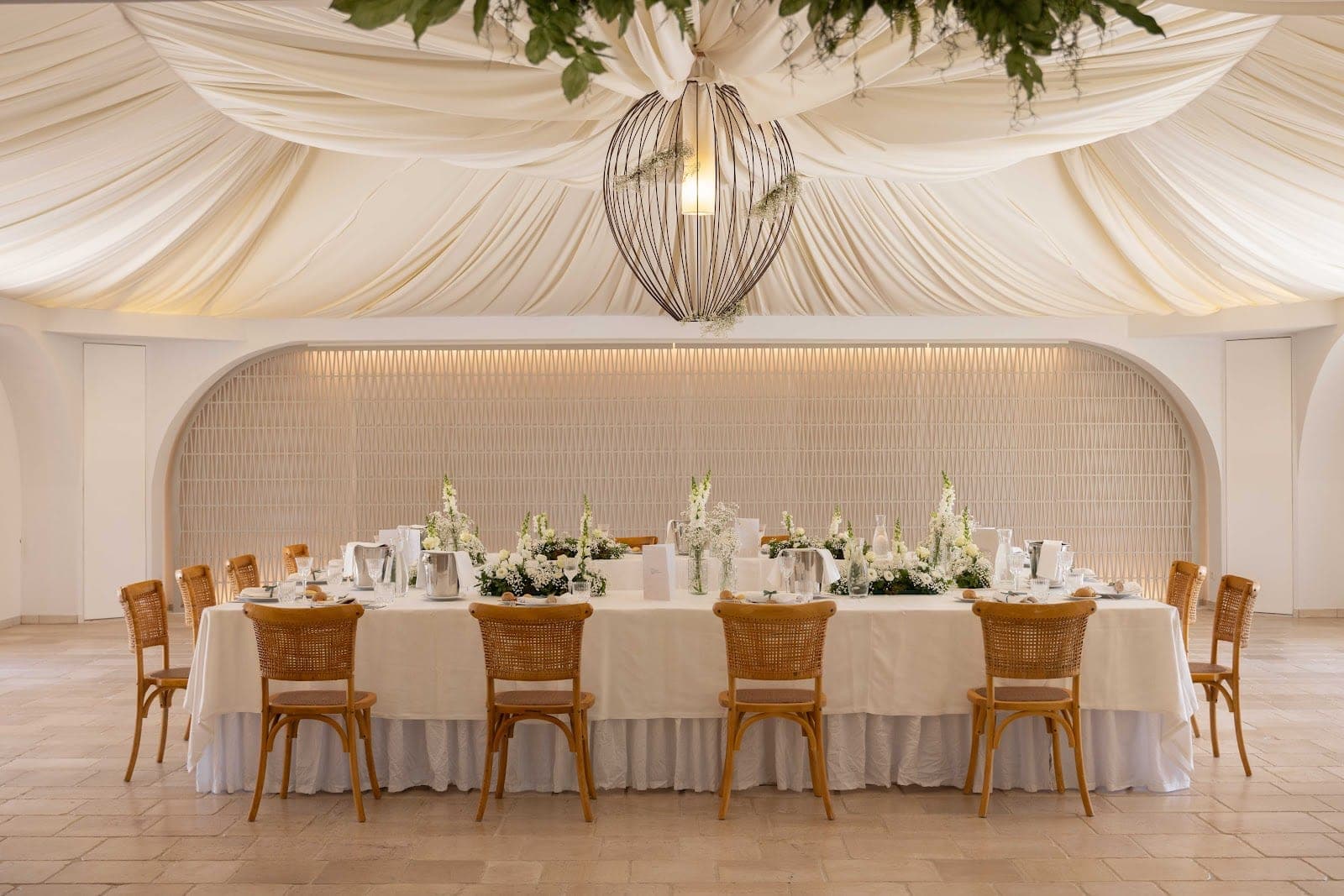 A long reception table set with white linens, floral centerpieces, and chairs.