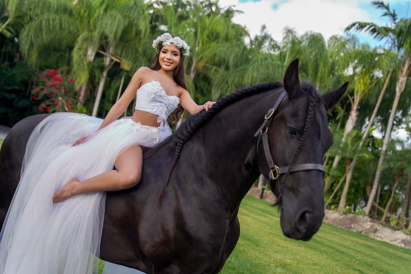A young woman in a white dress sits on a black horse in a garden.