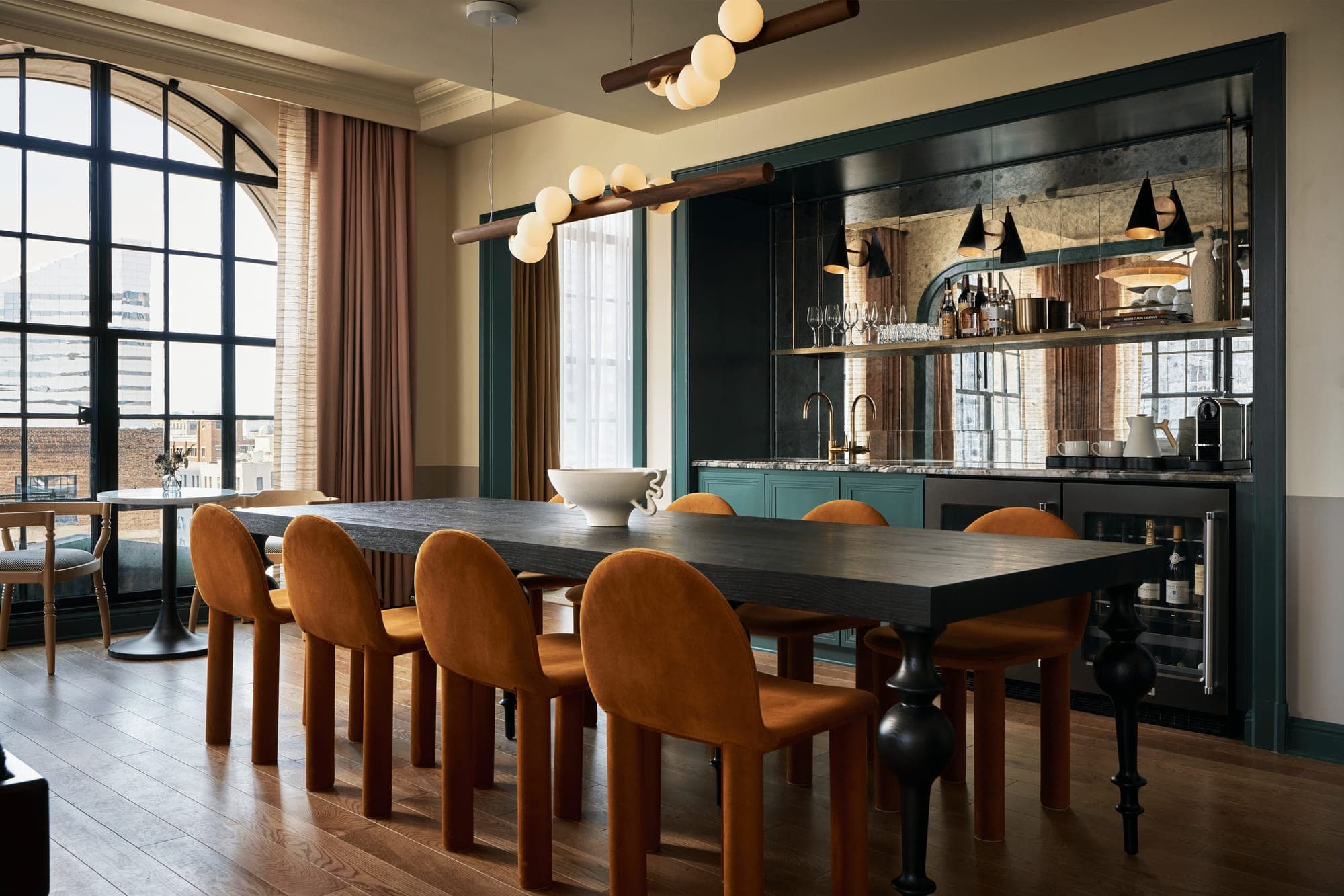 A long dark dining table with orange chairs in a room with a large window and a bar area.