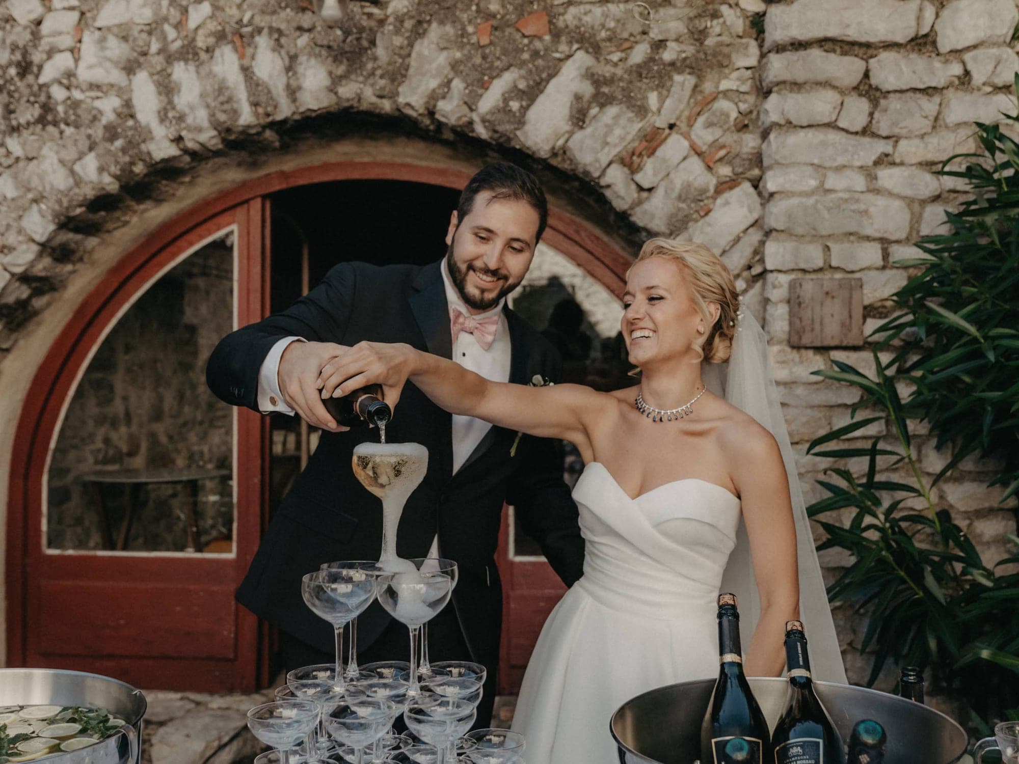 A bride and groom pour champagne into a pyramid of glasses.