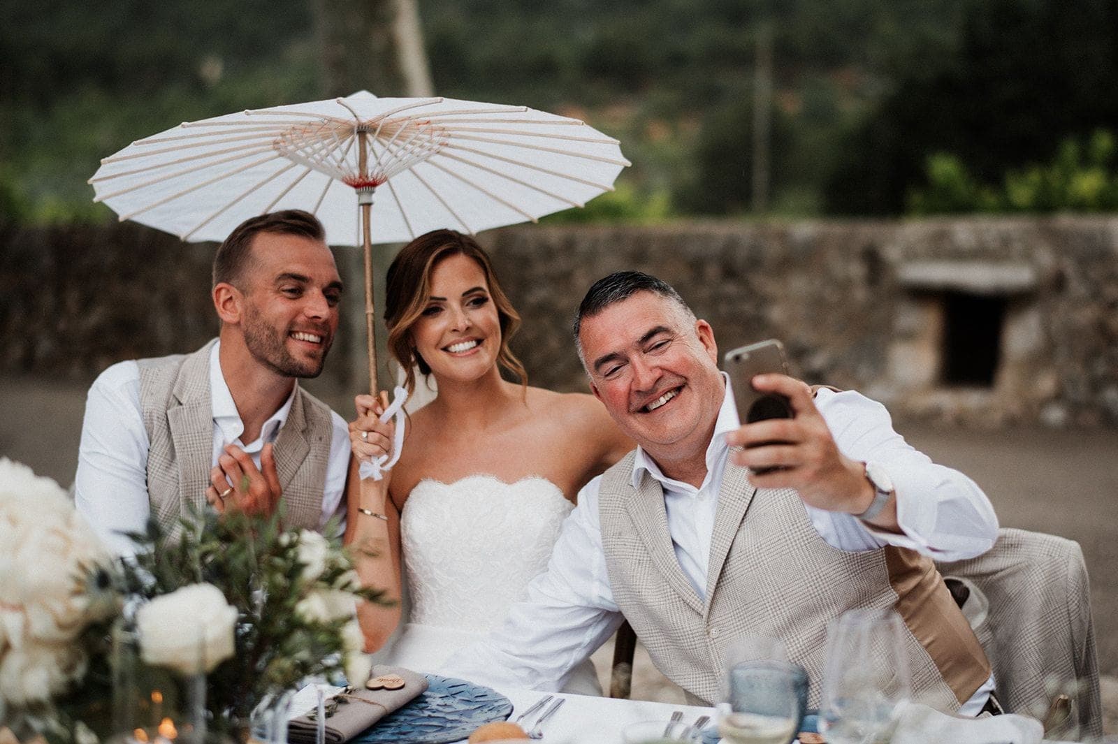A bride, groom, and another man take a selfie at an outdoor wedding reception table.