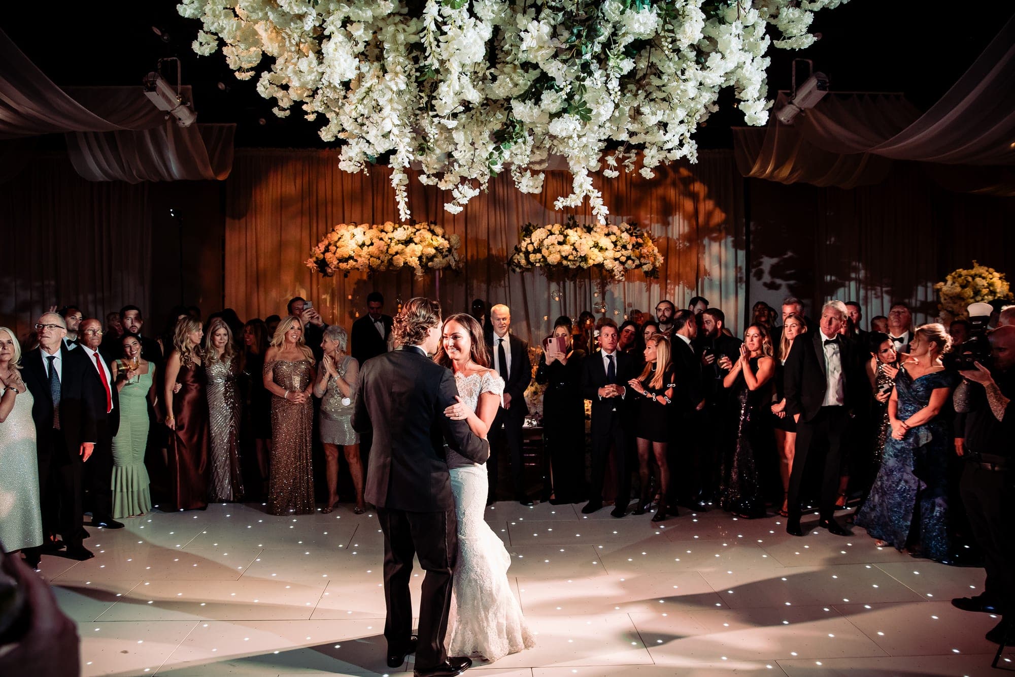 A couple dances on a lit dance floor surrounded by wedding guests.