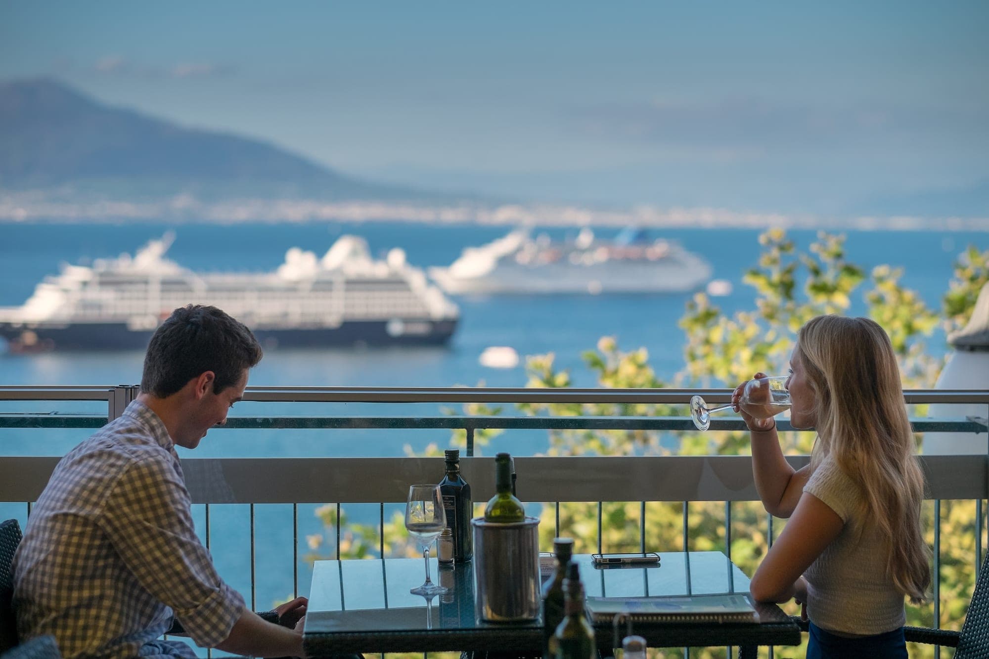 A couple sits at a table on a terrace overlooking the sea with cruise ships in the distance.