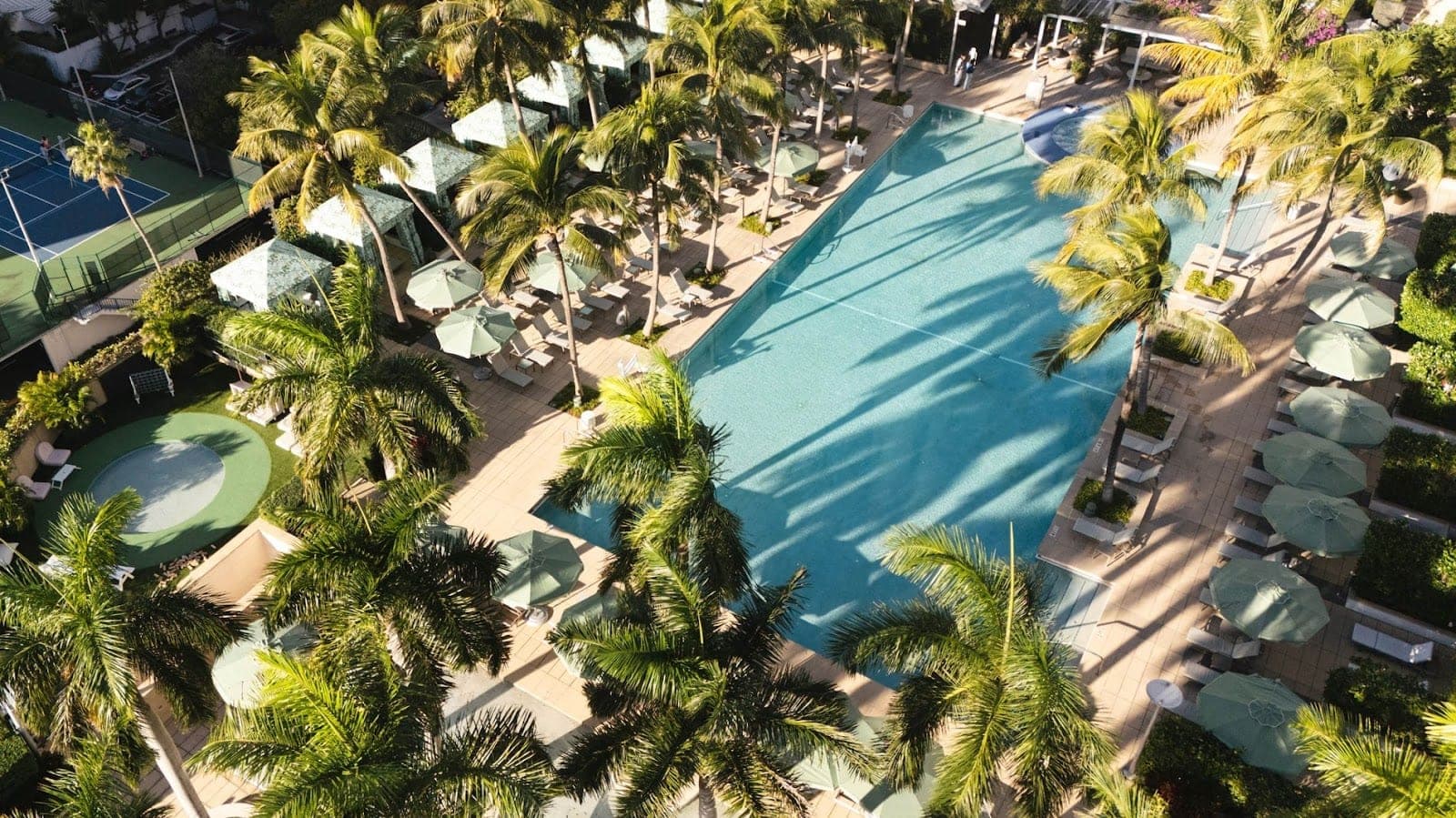 Aerial view of a resort pool and tennis courts surrounded by palm trees.