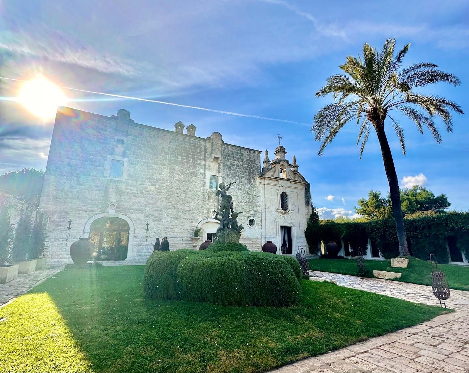 Exterior of a stone building with a palm tree and a statue under a bright blue sky.