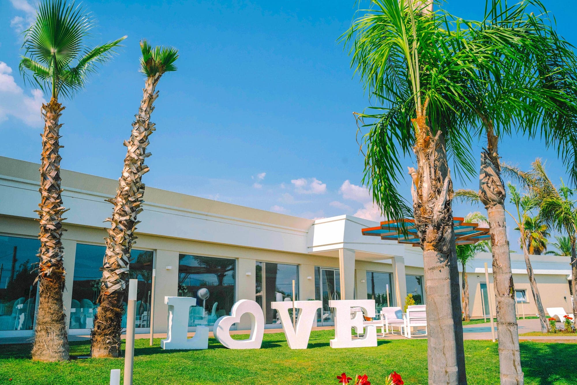 Large white LOVE letters on a green lawn in front of a white building with palm trees.