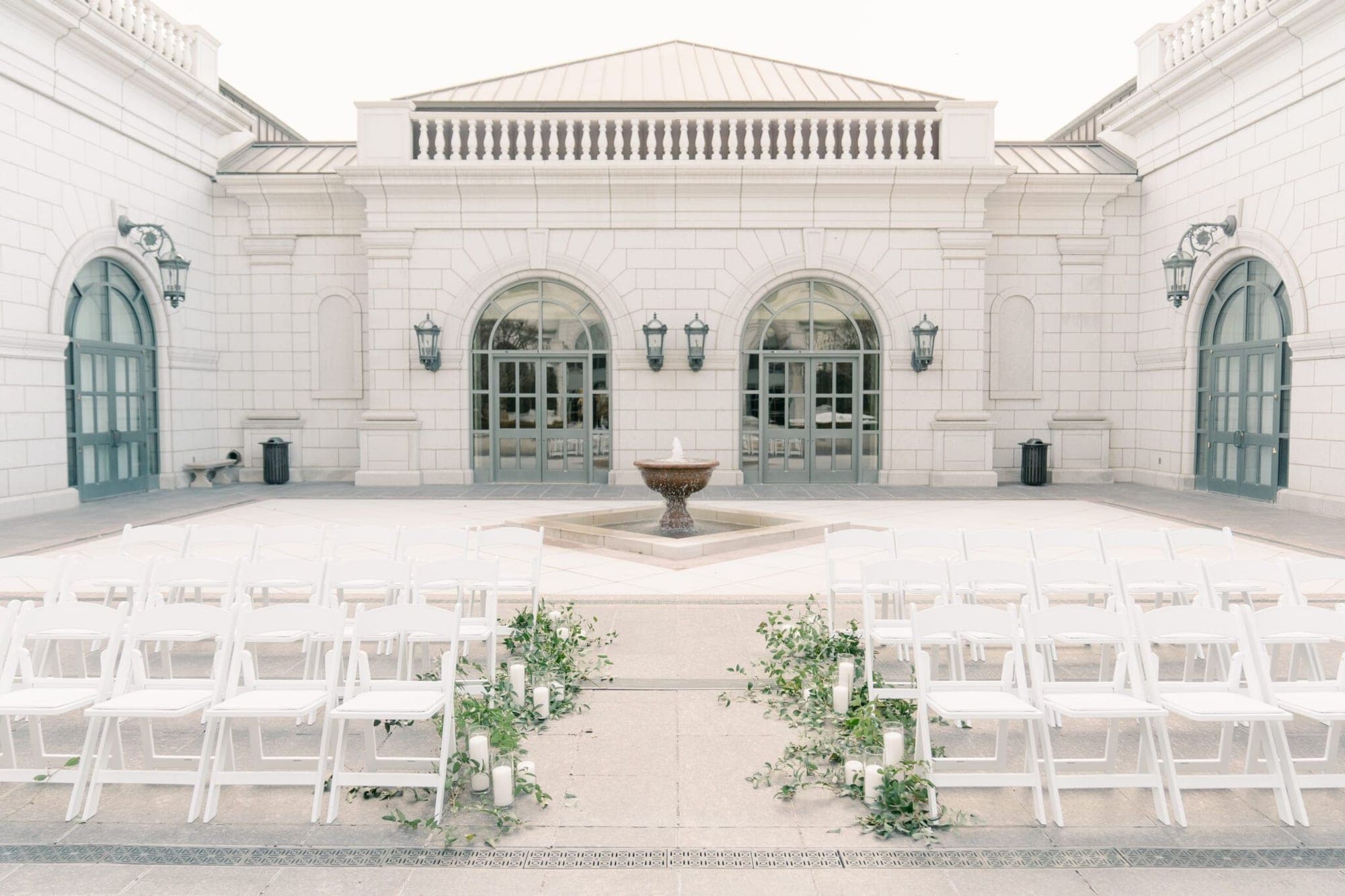 Wedding ceremony setup with white chairs and a fountain in front of a building.