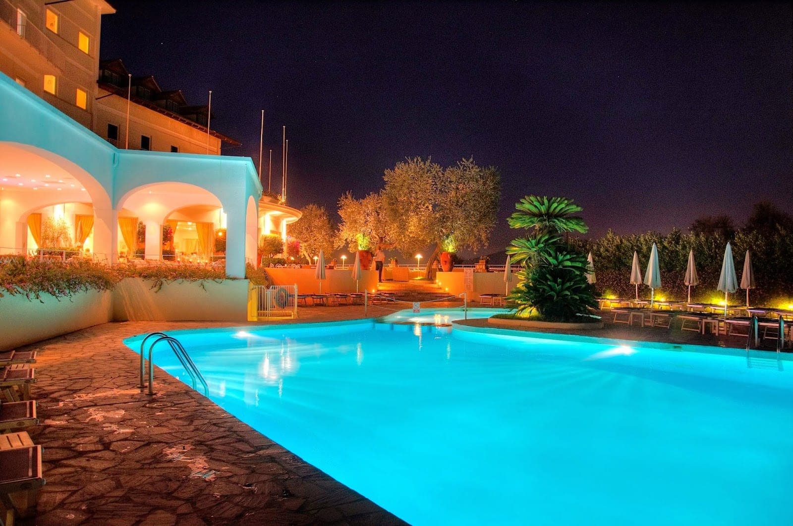 A brightly lit swimming pool at night with lounge chairs and umbrellas around it.