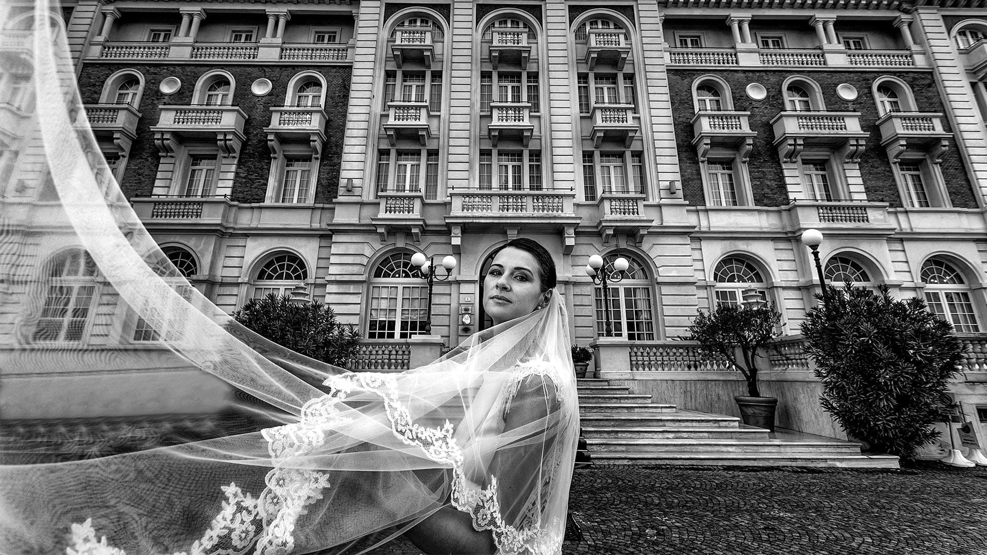 Bride with veil blowing in the wind in front of a large, ornate building.