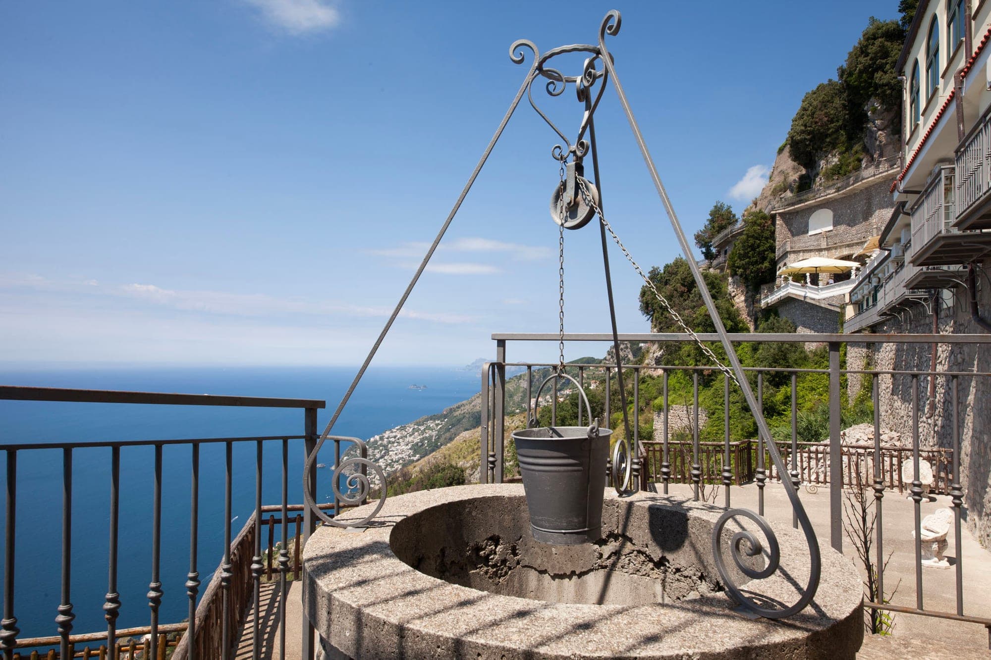 A well with a bucket and pulley system overlooks the sea and a distant town.