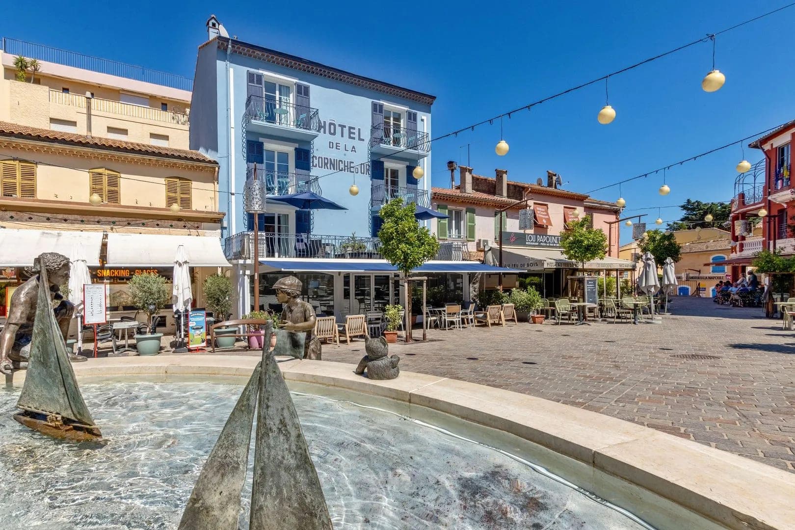 Exterior of Hotel de la Corniche d'Or with outdoor seating and a fountain.