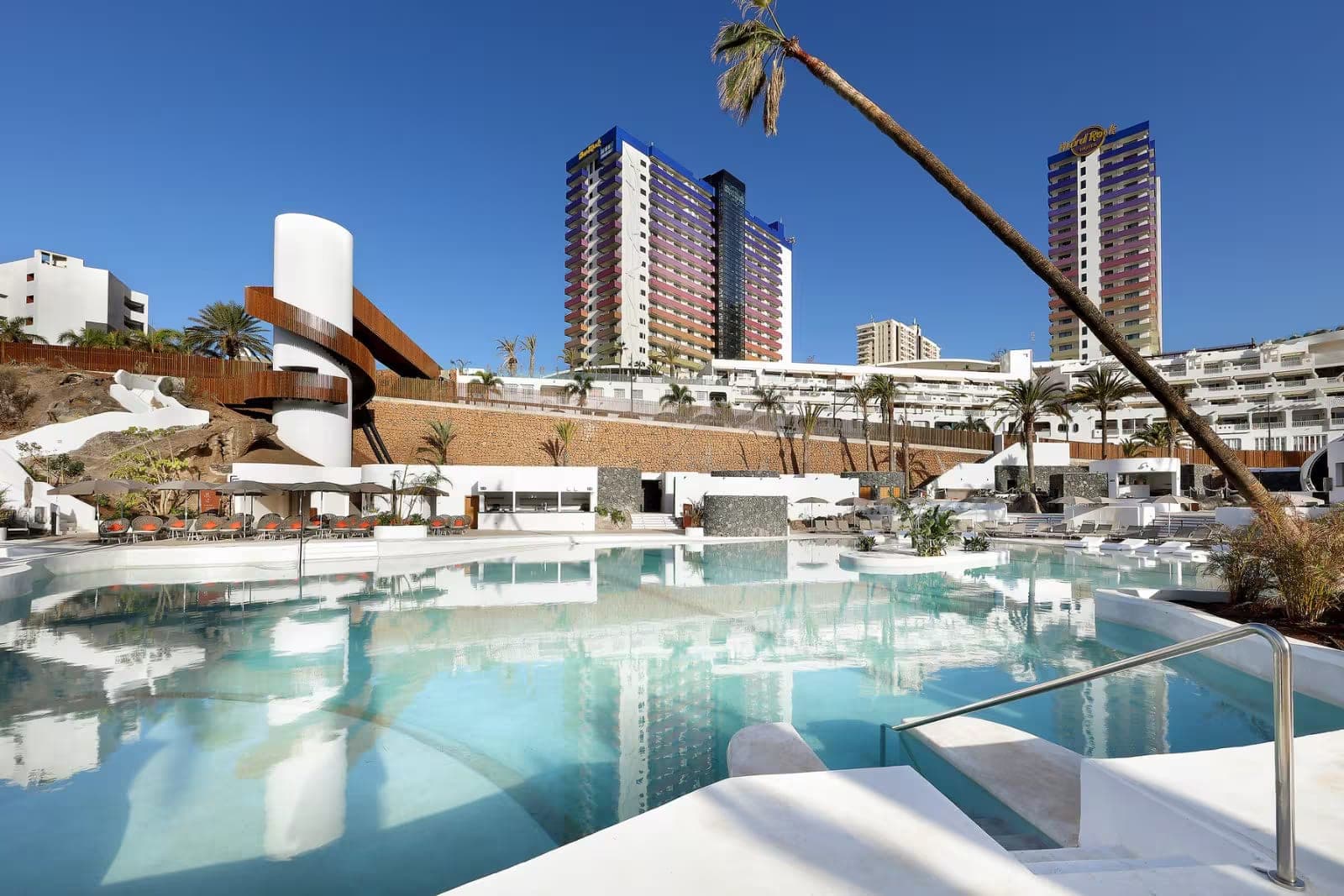 A swimming pool with lounge chairs and palm trees in front of hotel buildings.