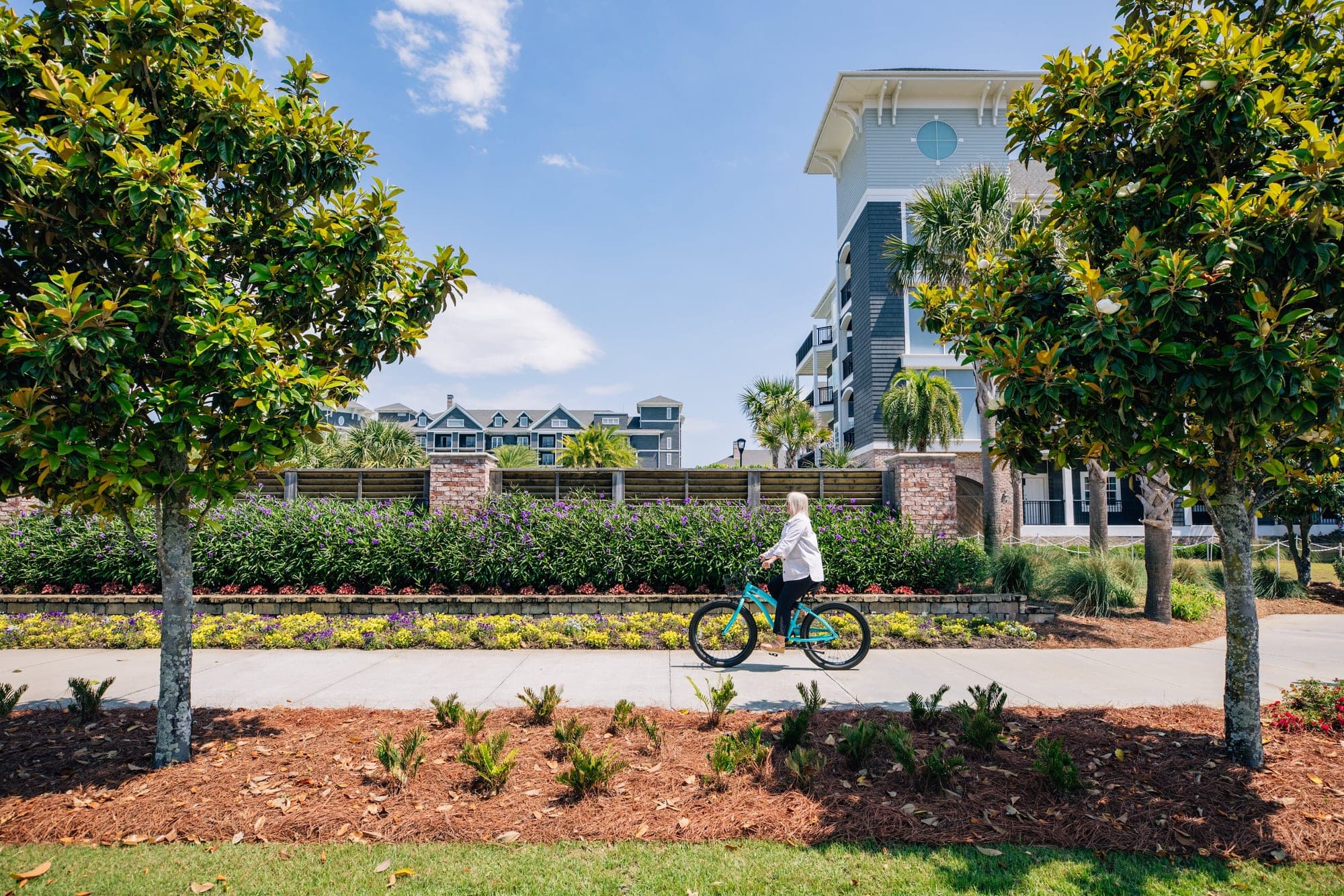 A woman rides a bicycle on a path with buildings and trees.