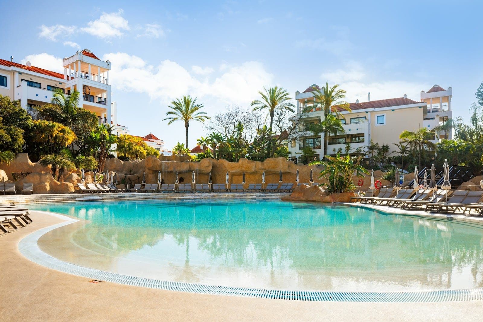 Swimming pool with lounge chairs and resort buildings under a blue sky.