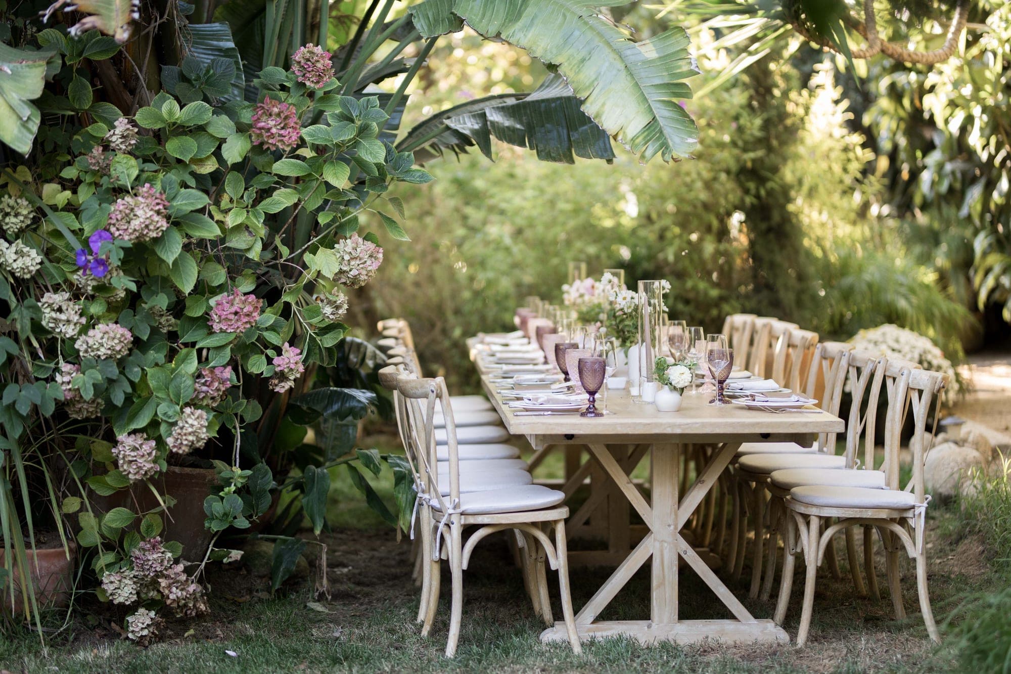 A long wooden table set for a reception in a garden with chairs and floral centerpieces.