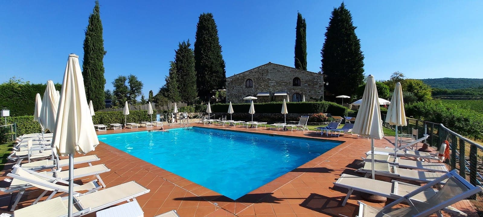 Swimming pool area with lounge chairs and umbrellas, overlooking vineyards and a stone building.