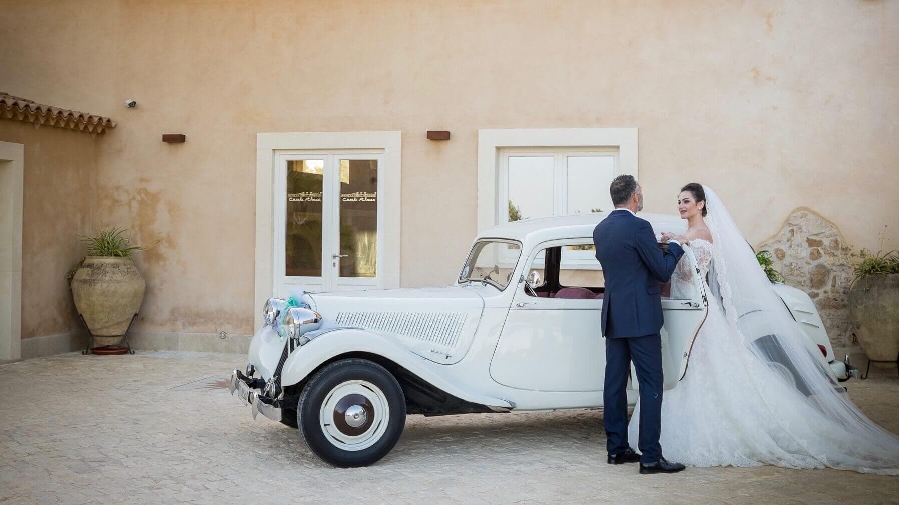 Bride and groom stand by a white vintage car outside a building.