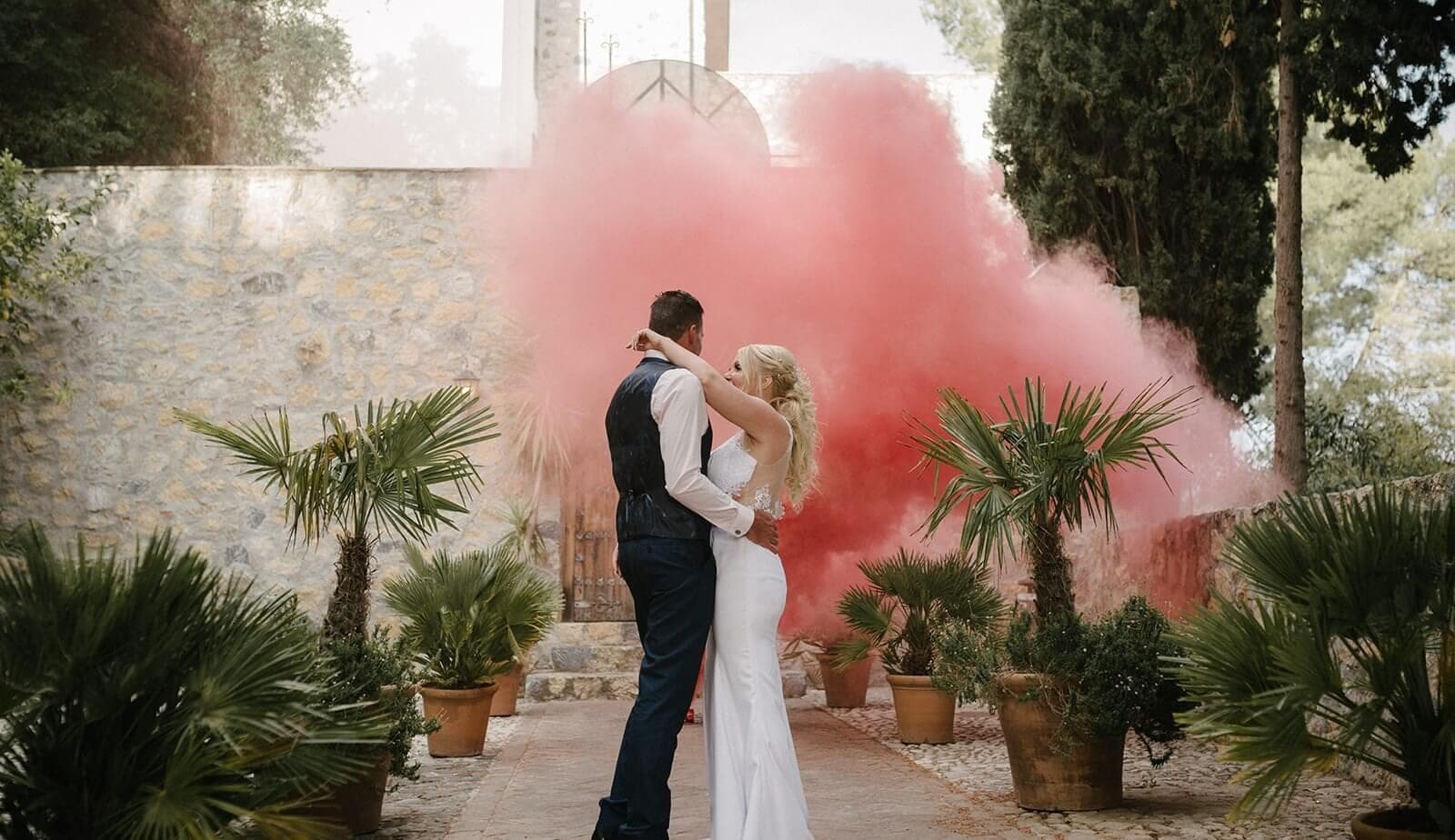 A bride and groom embrace in a courtyard with pink smoke behind them.