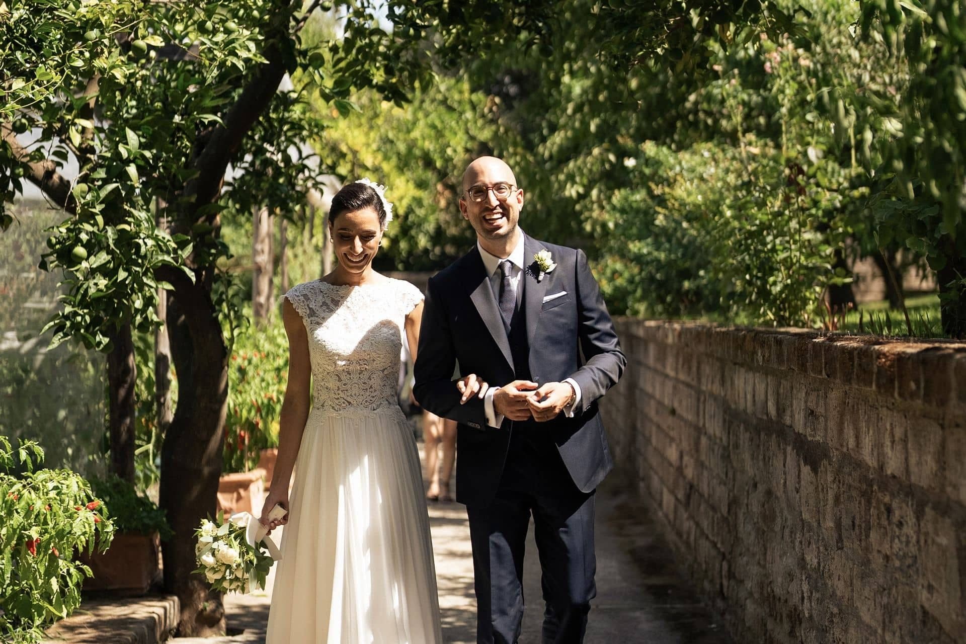 A smiling bride and groom walk down a path in a garden.