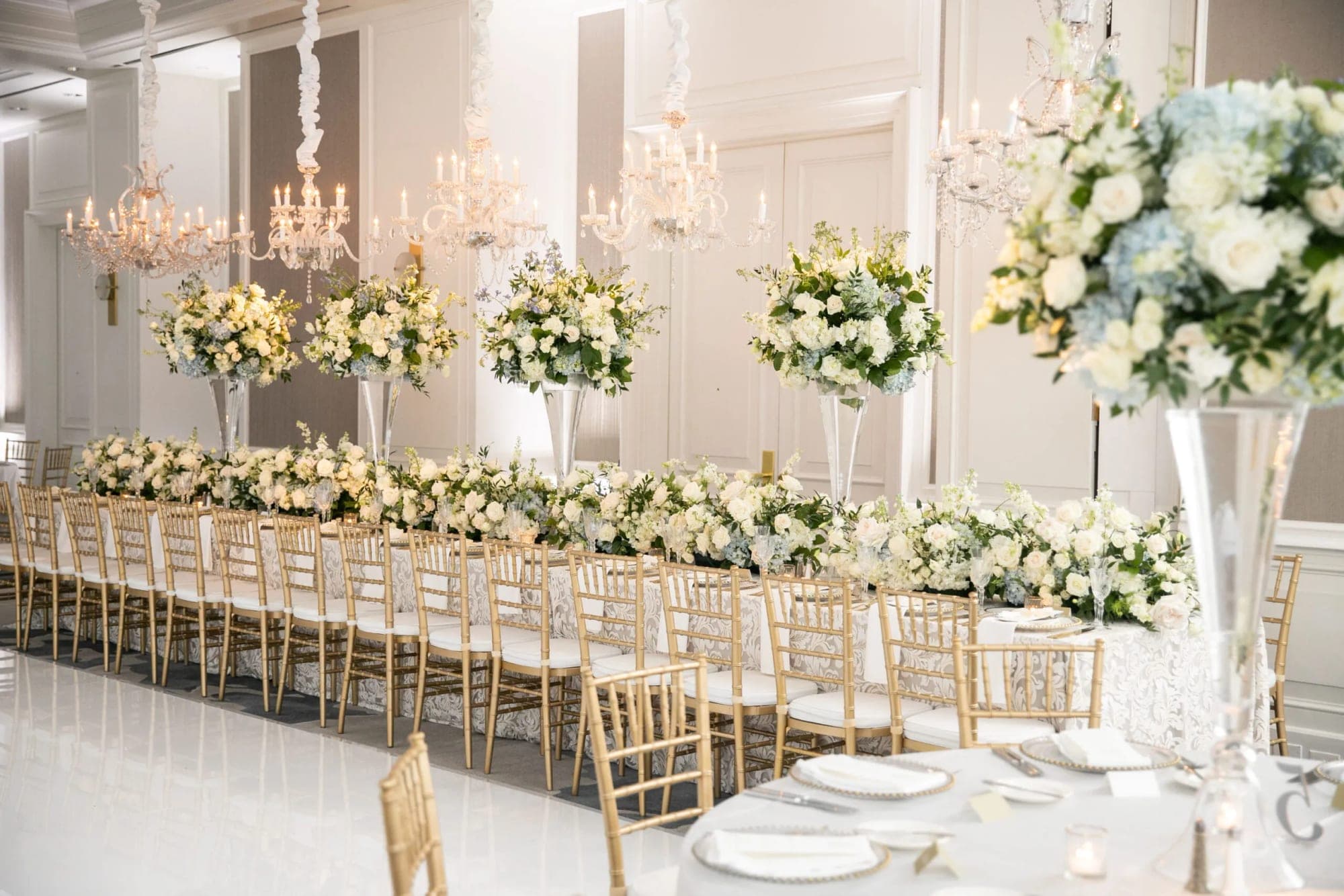 A long reception table set with white linens, gold chairs, and abundant white and blue floral arrangements.