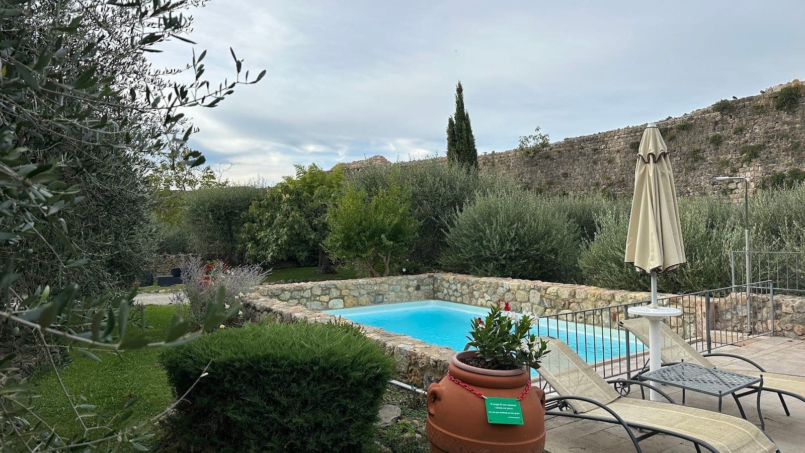 A swimming pool area with lounge chairs and an umbrella, set against a stone wall and olive trees.