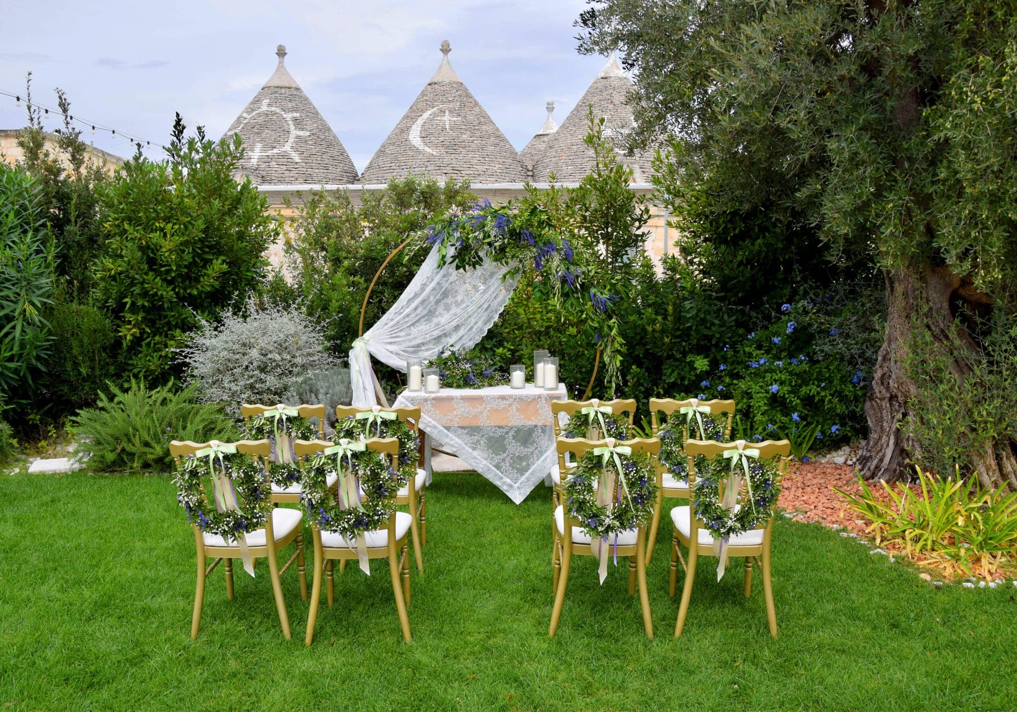 Wedding ceremony setup with chairs and an altar in a garden with trulli buildings.