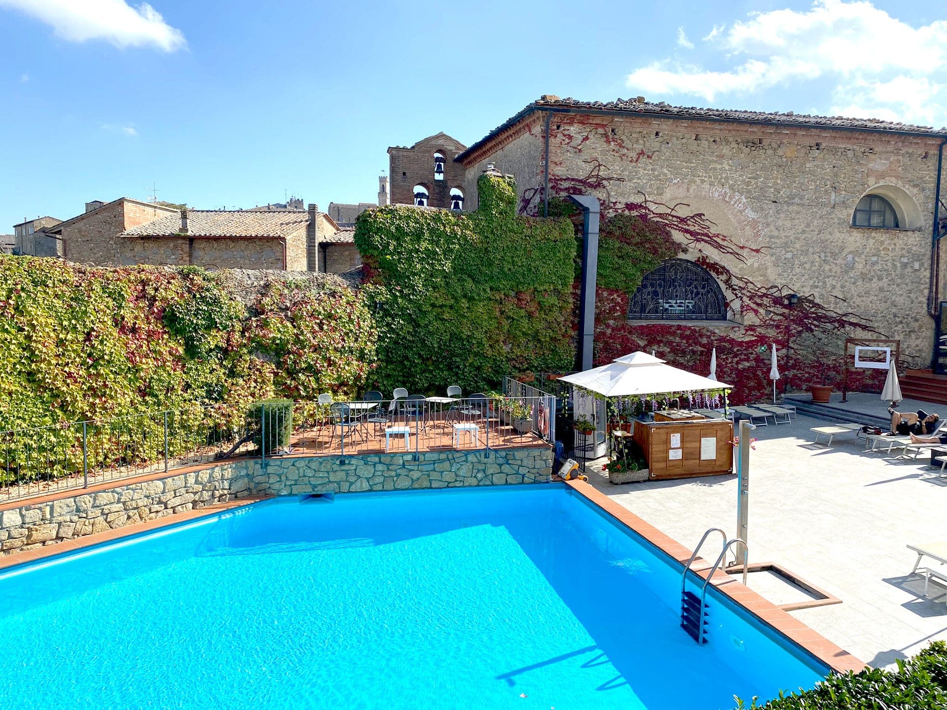 A bright outdoor swimming pool area with lounge chairs and a building covered in ivy.