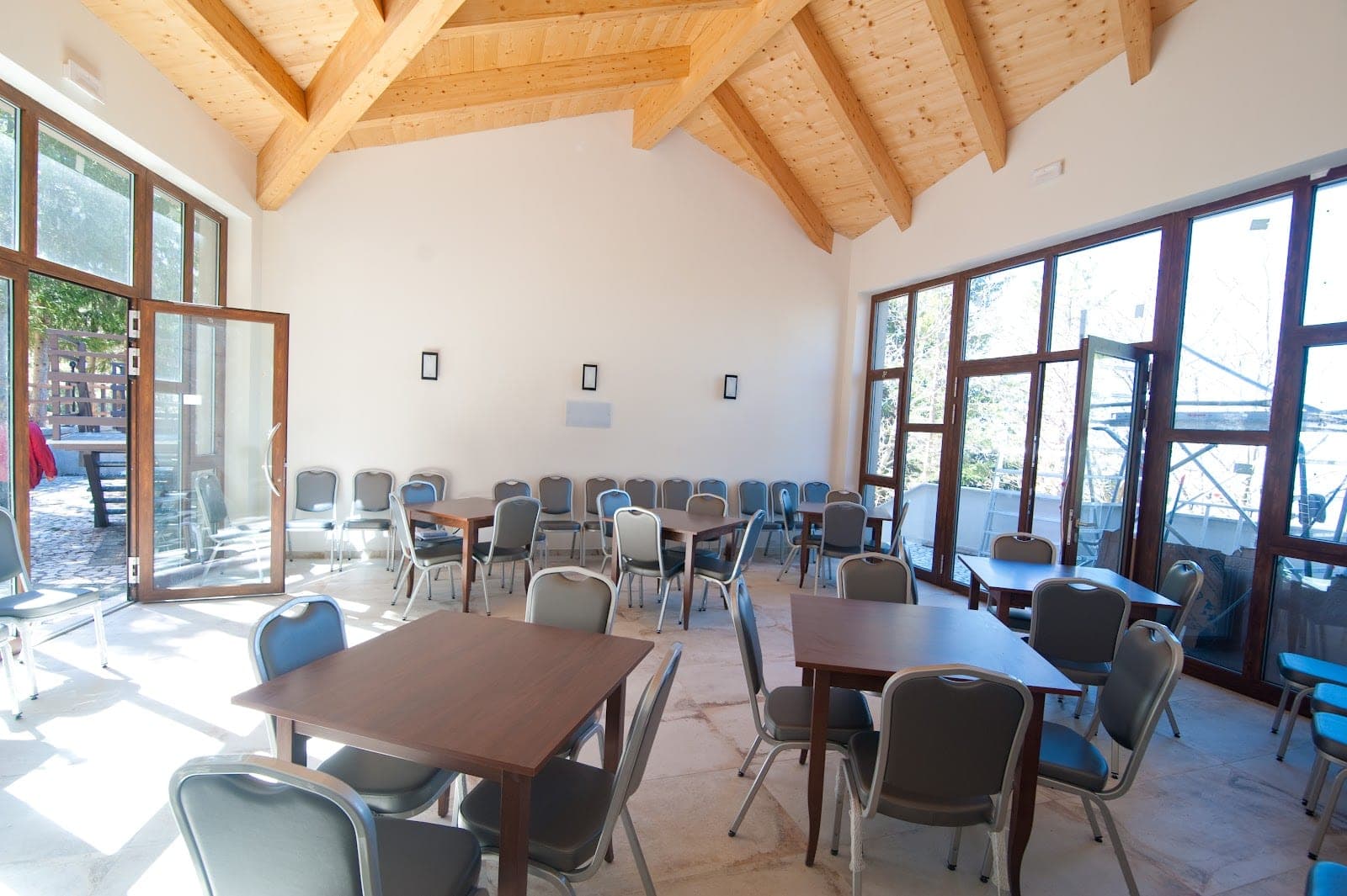 A room with tables and chairs, large windows, and wooden ceiling beams.