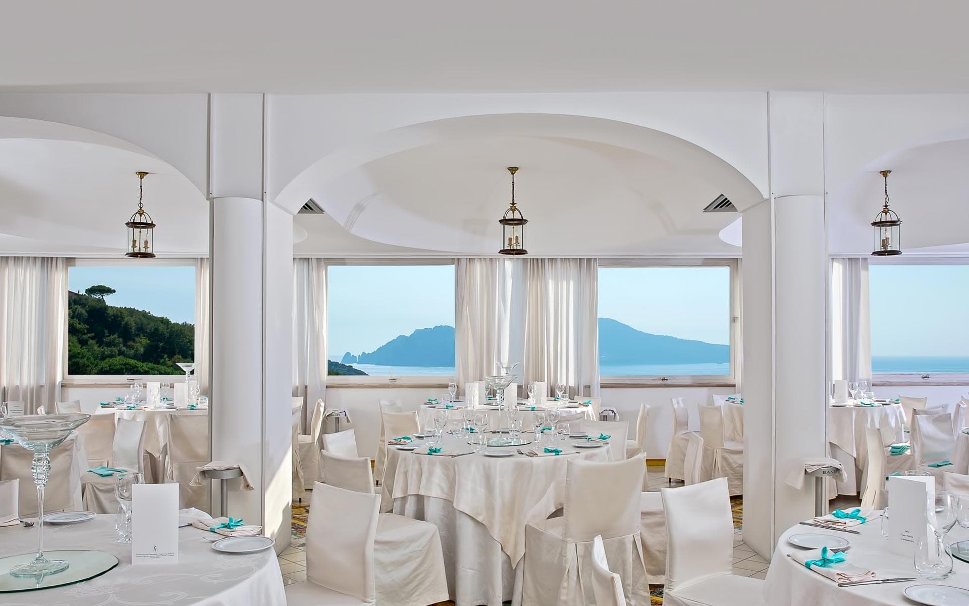 A wedding reception dining room with tables set for guests and large windows overlooking the sea and mountains.