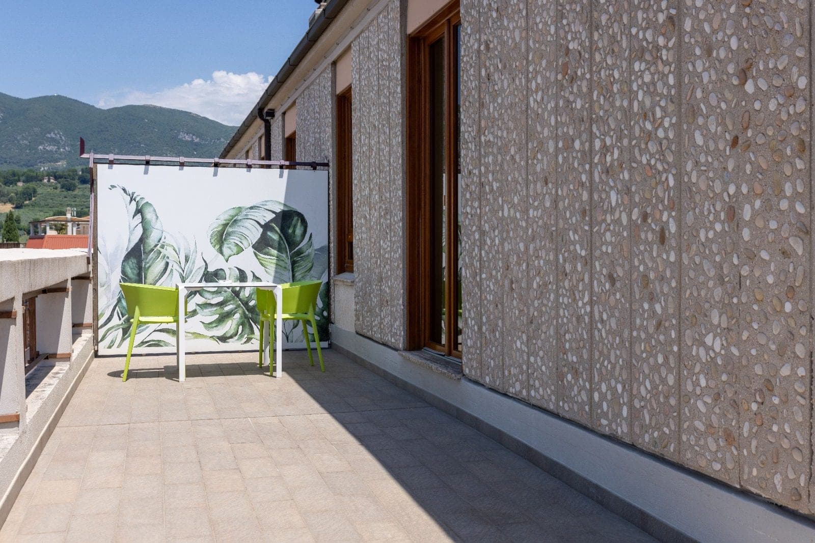 Balcony with two green chairs and a table against a tropical leaf backdrop.