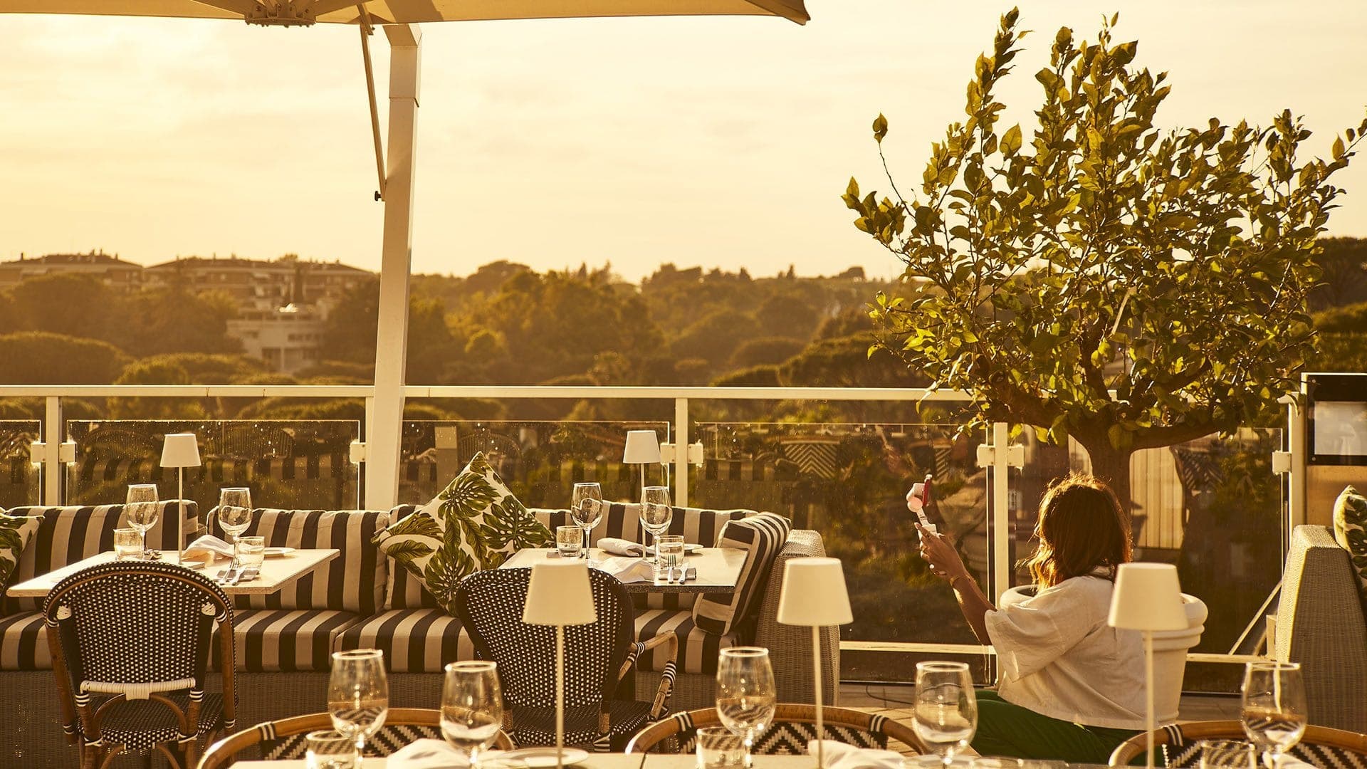 A terrace with tables and seating overlooking a city and trees at sunset.