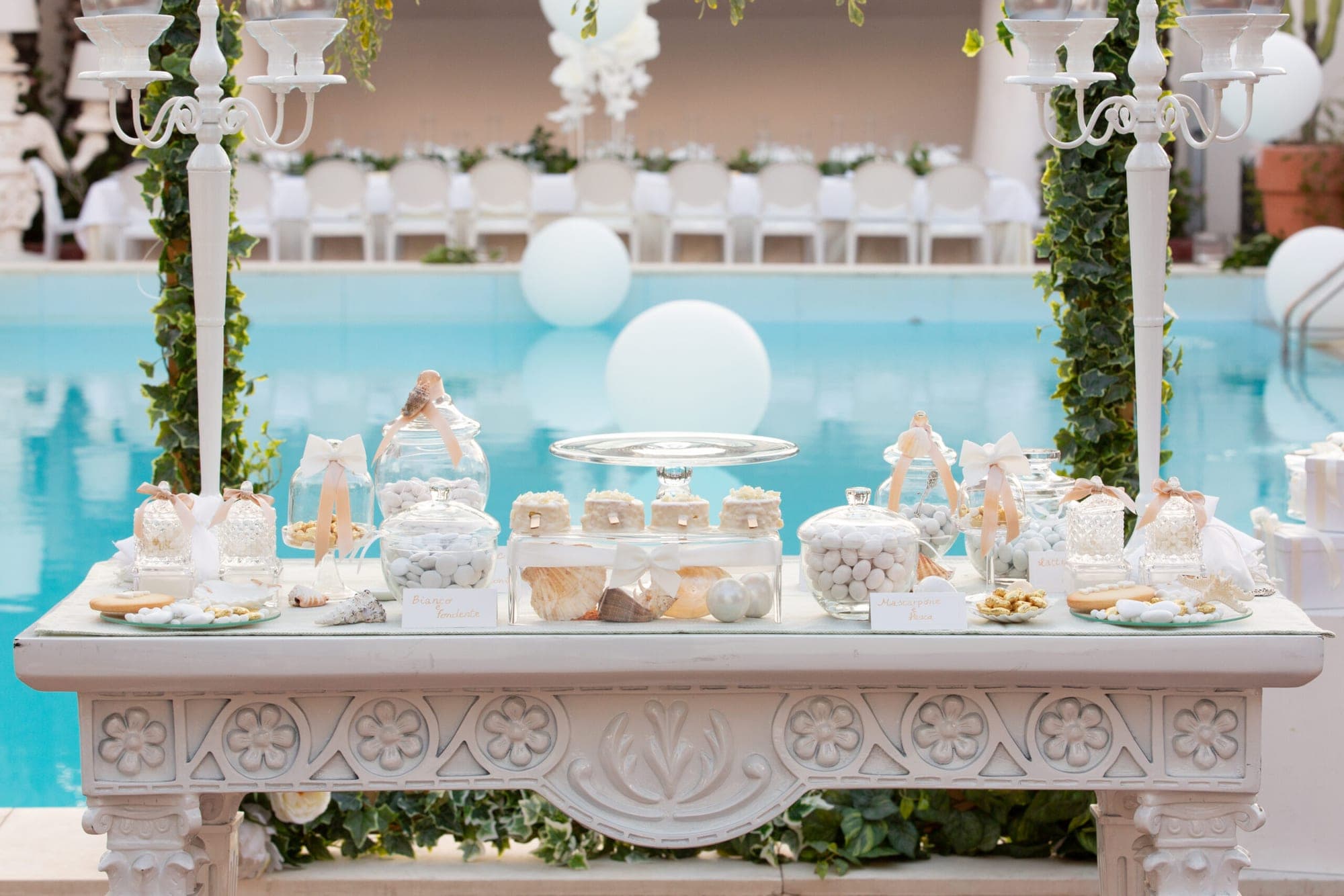 Dessert table with candies and cakes by a swimming pool with chairs in the background.