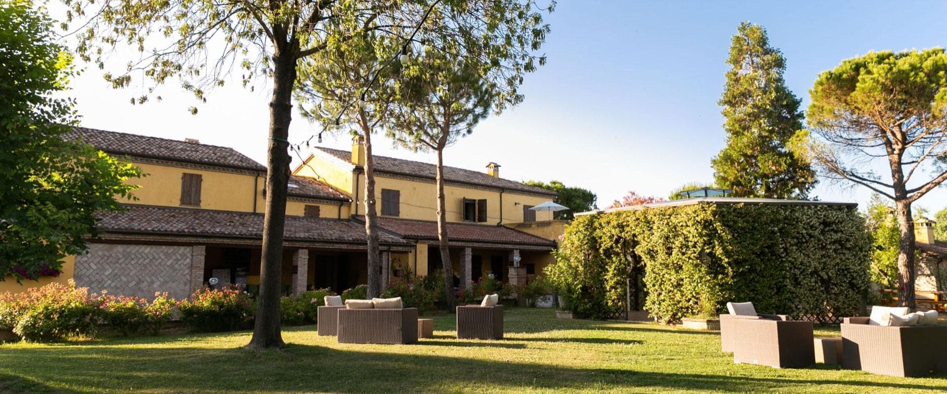 Exterior of a yellow building with a tiled roof, surrounded by trees and a lawn with outdoor furniture.