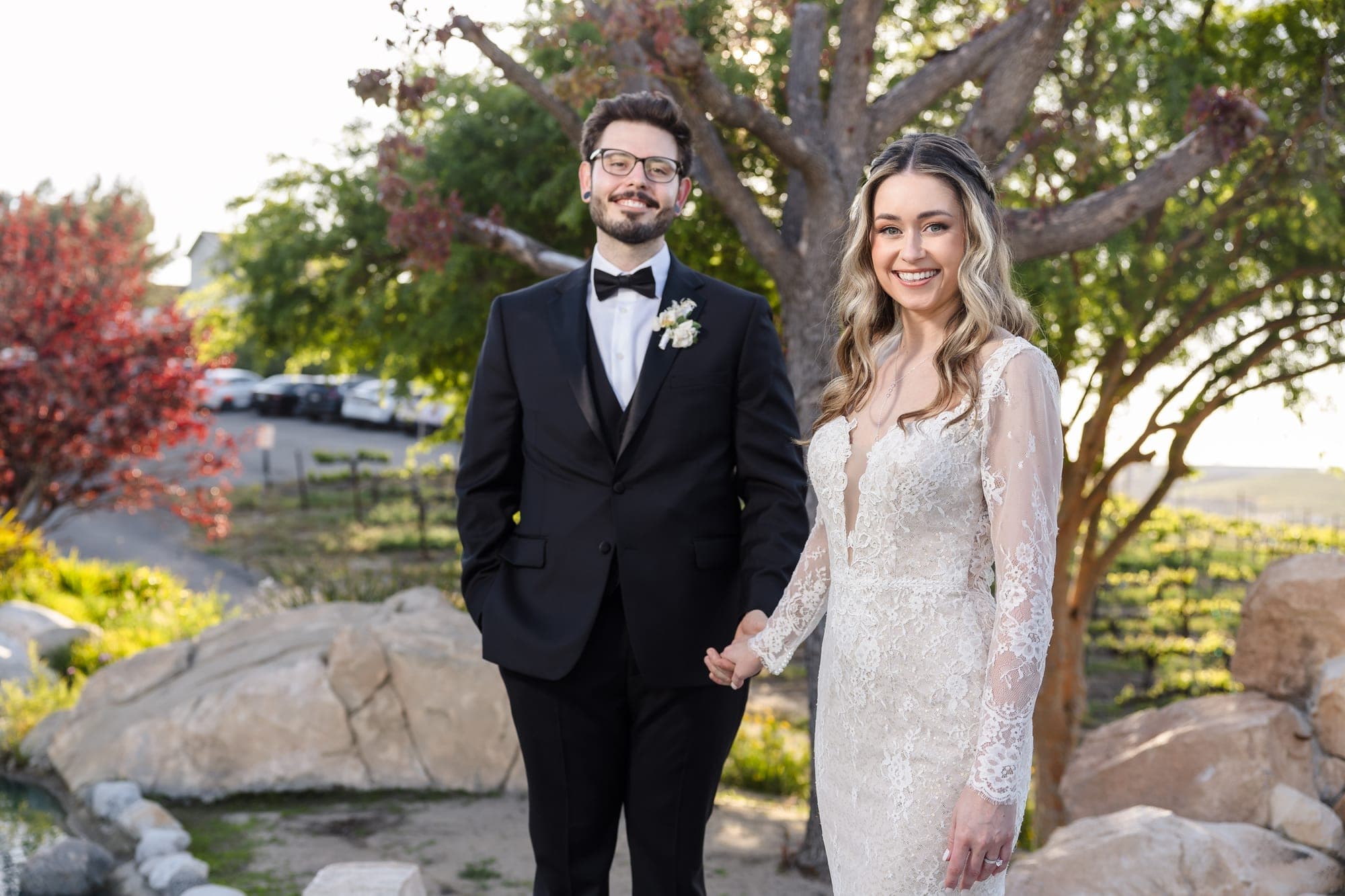 Bride and groom holding hands outdoors with vineyard in background.