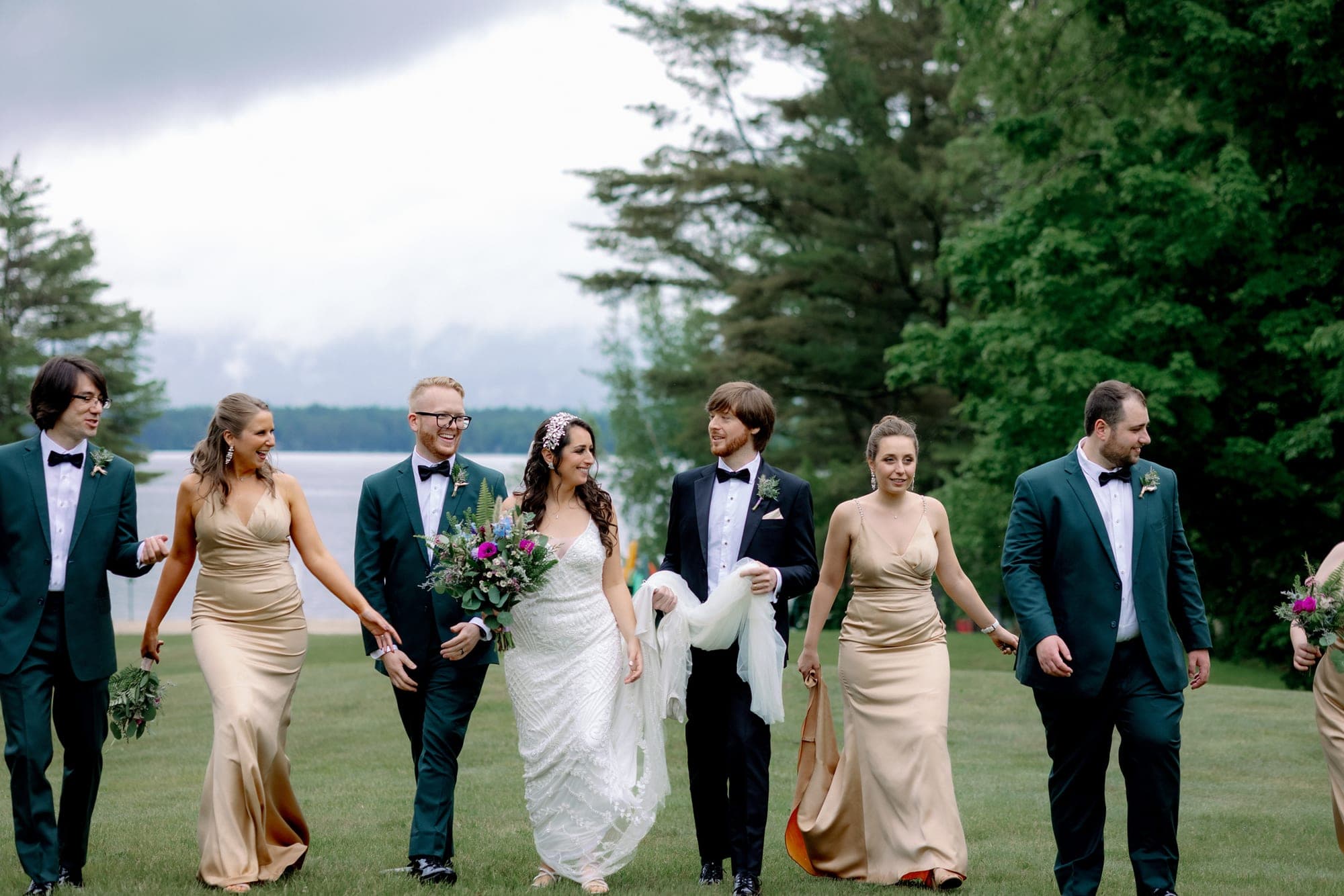 A wedding party walks across a grassy area with a lake and trees in the background.