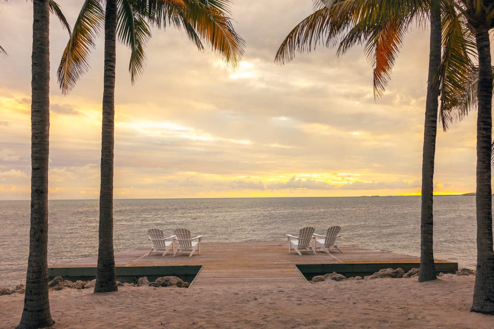 Wooden deck with chairs facing the ocean at sunset, framed by palm trees.