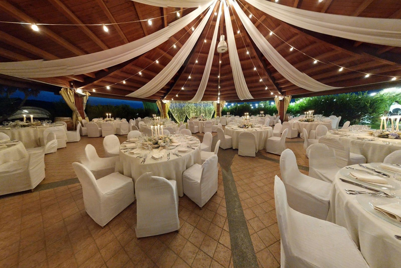 Outdoor reception area with round tables covered in white cloths and chairs with white covers.