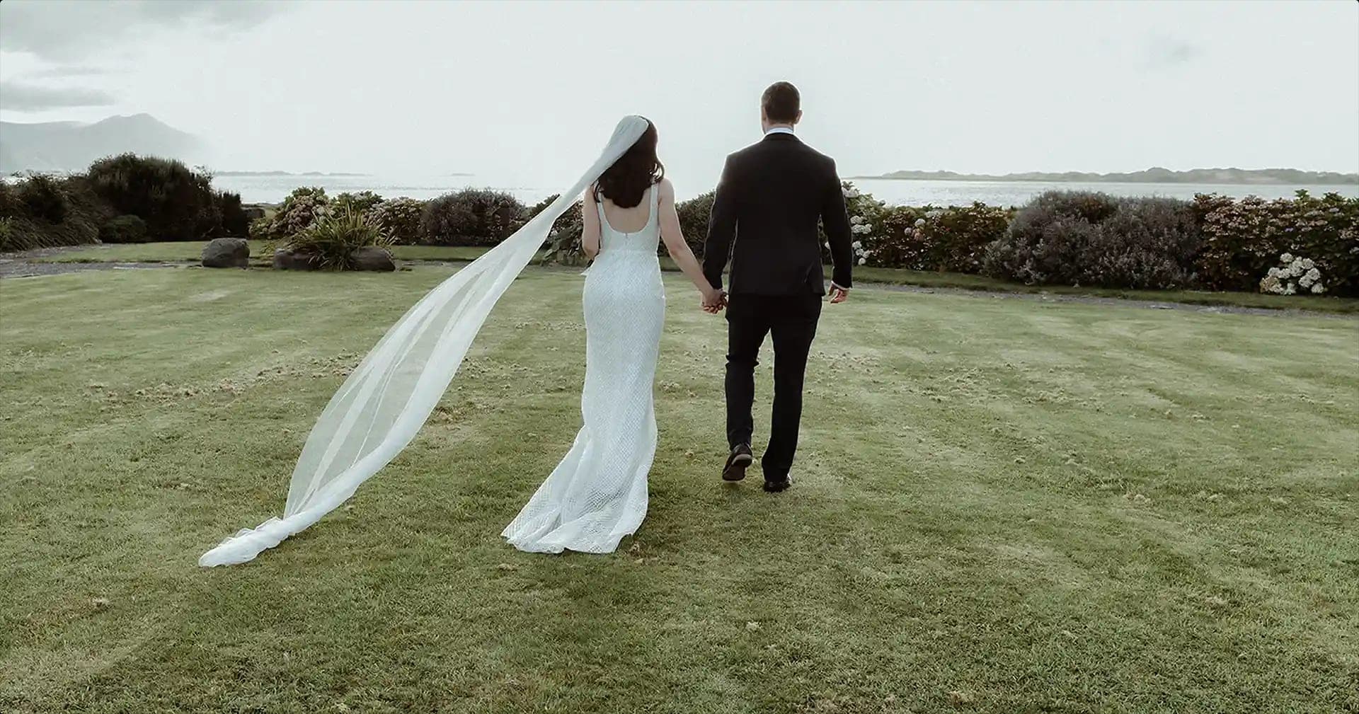 Bride and groom walking on grass towards the sea.