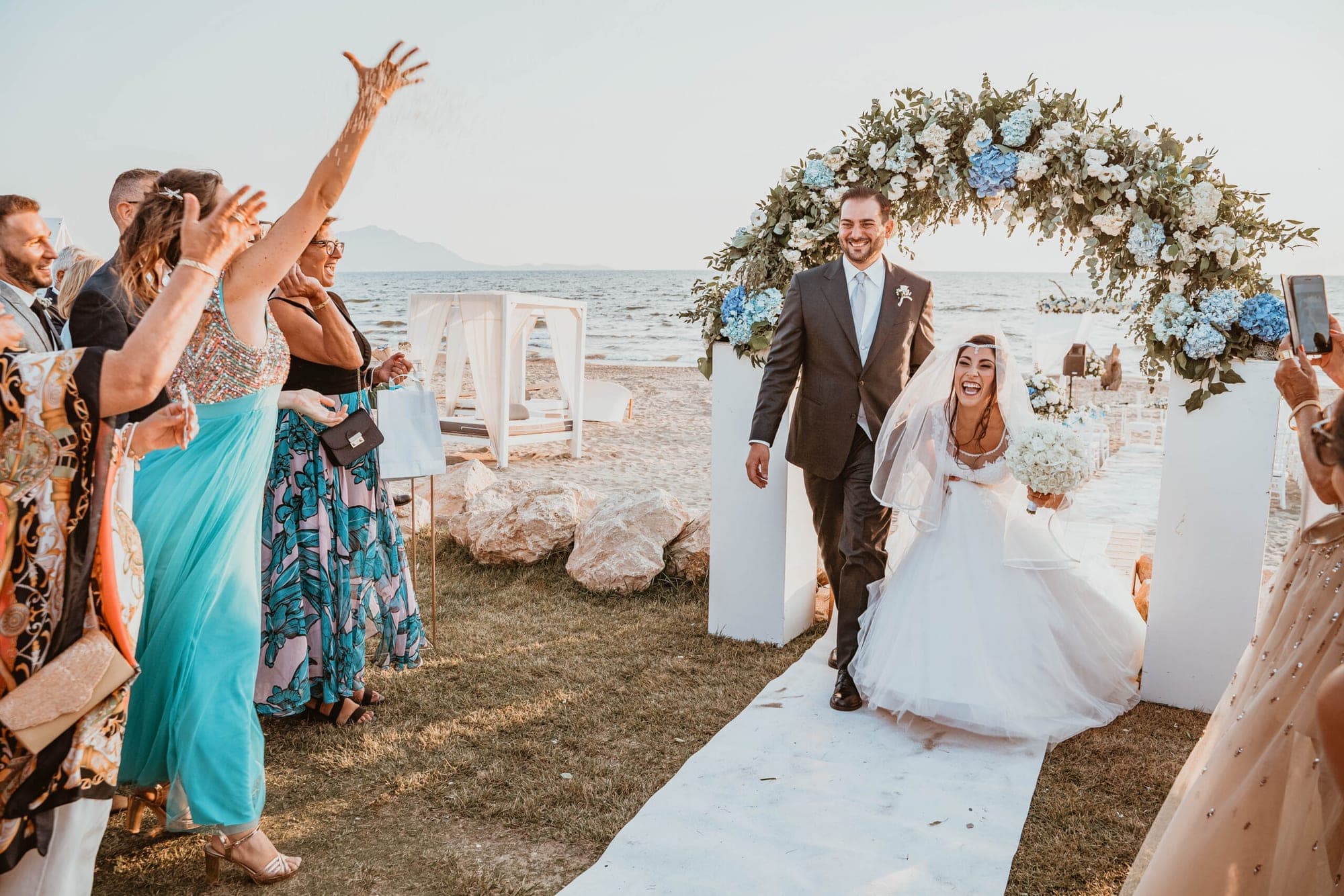 Newlyweds walk down a path towards the sea with guests.
