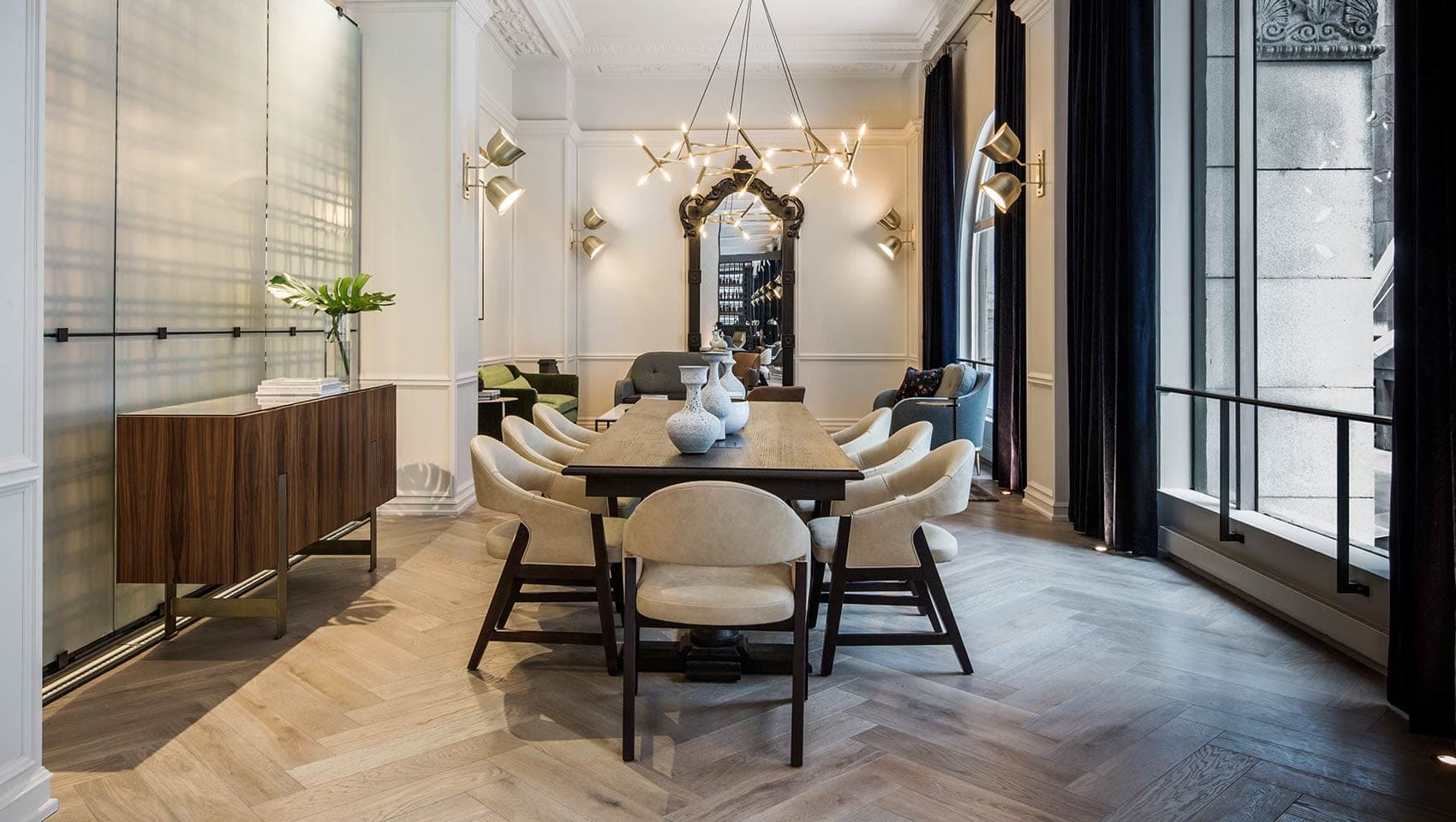 A long dining table with chairs is set in a room with a large mirror and chandelier.